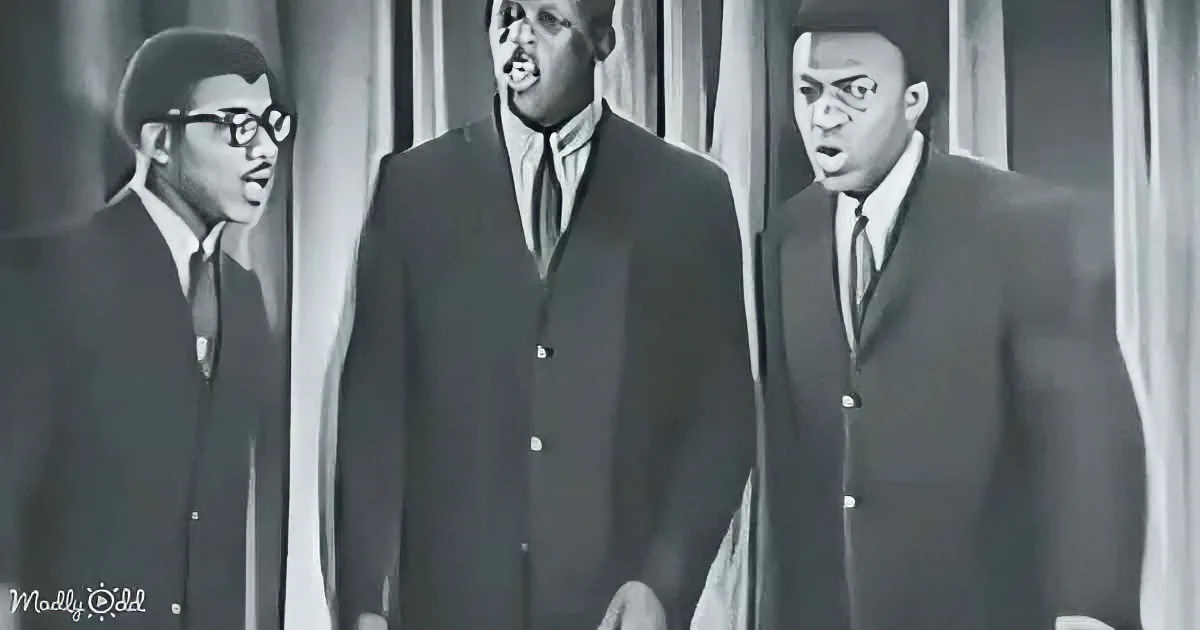 Three men in suits sing together, one wearing glasses, against a striped curtain backdrop.