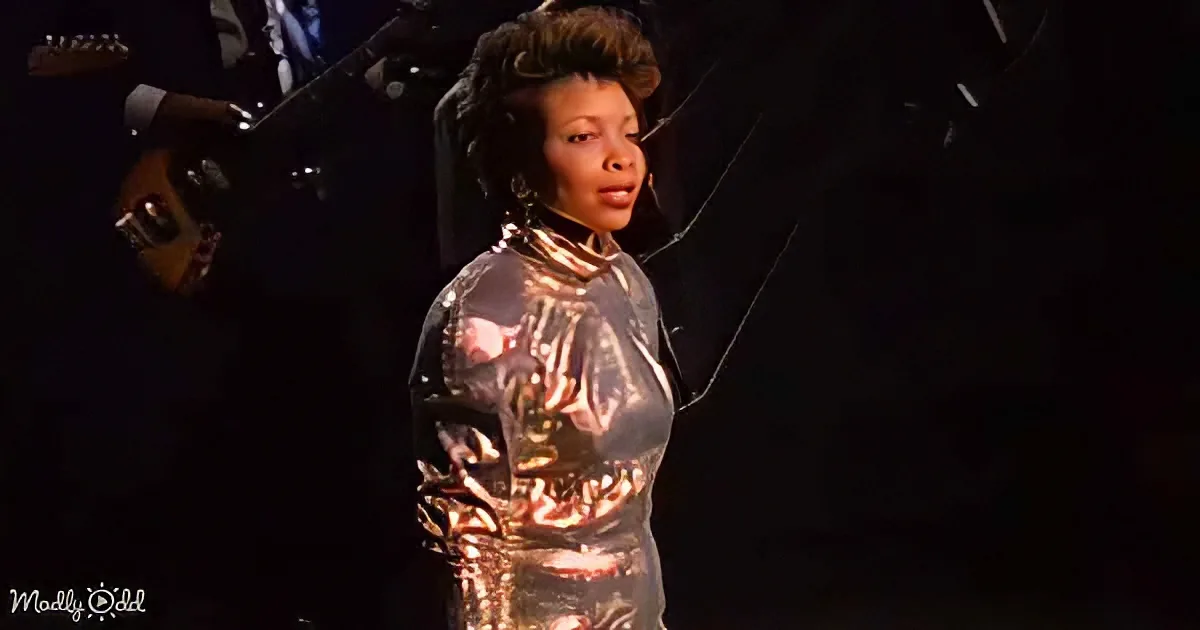 A performer in a shimmering silver outfit sings on stage, surrounded by musicians.