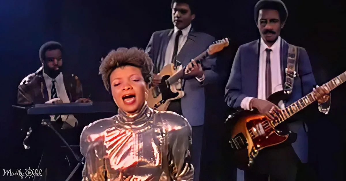 A performer in a shiny outfit sings passionately, backed by a band in suits playing instruments.