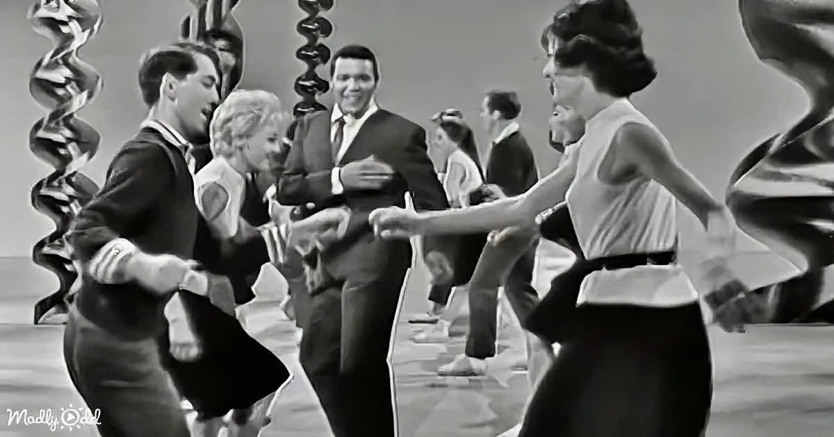A man in a suit dances joyfully with a woman in a white blouse, surrounded by other couples in a lively dance scene.
