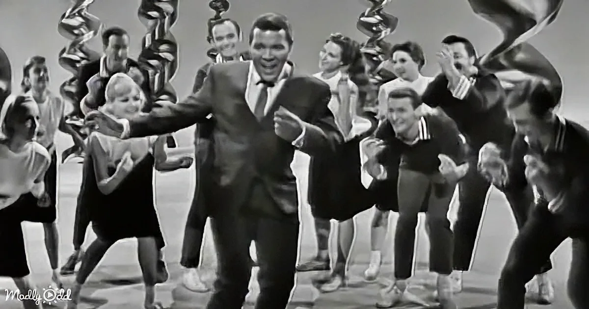 A man in a suit dances joyfully in front of a group of people, all smiling and clapping.