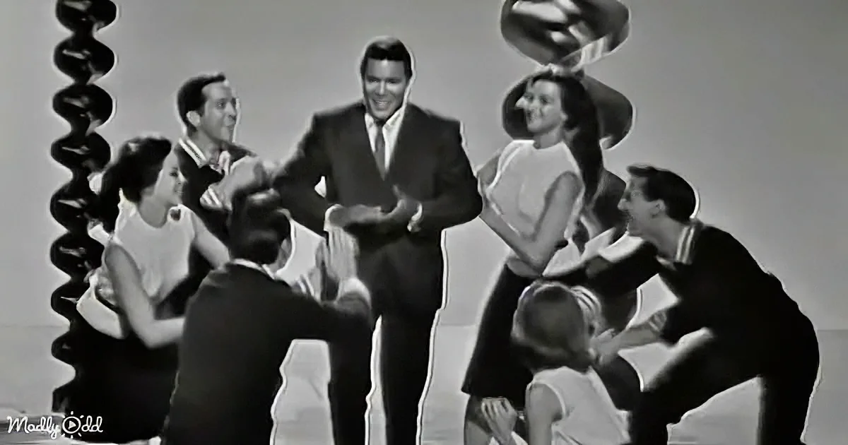 A man in a suit stands surrounded by dancers in a lively, vintage-style dance scene.