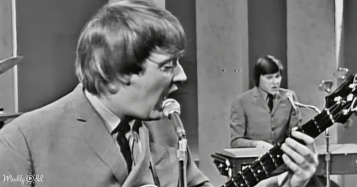 A man in a suit sings into a microphone, another person plays guitar in the background.