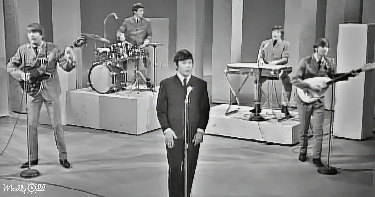 A man in a suit stands at a microphone, surrounded by musicians playing instruments in a studio setting.