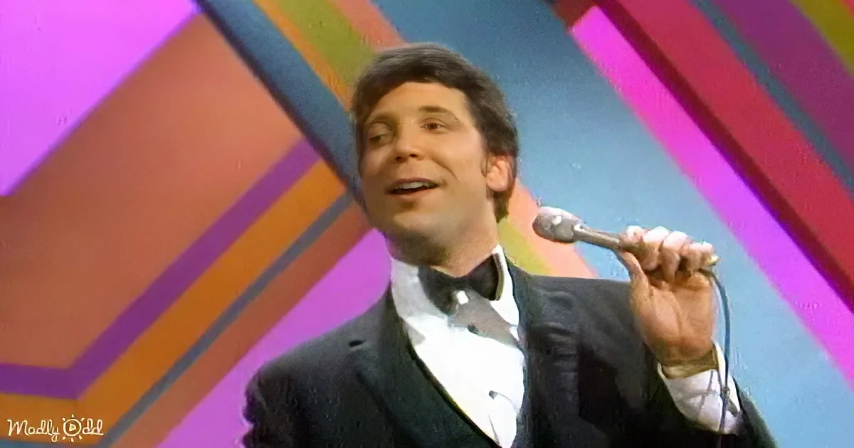 A man in a suit holds a microphone, smiling, against a colorful geometric background.