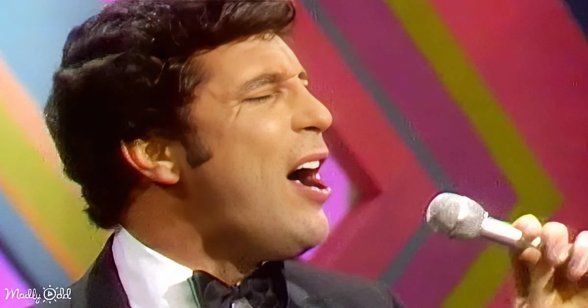 A man in a suit sings into a microphone, eyes closed, against a colorful background.