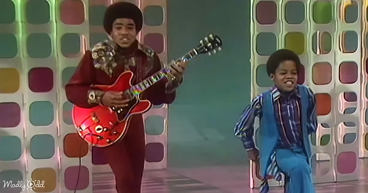 A man in a red suit plays a red guitar on stage, with another person in a blue outfit standing nearby.