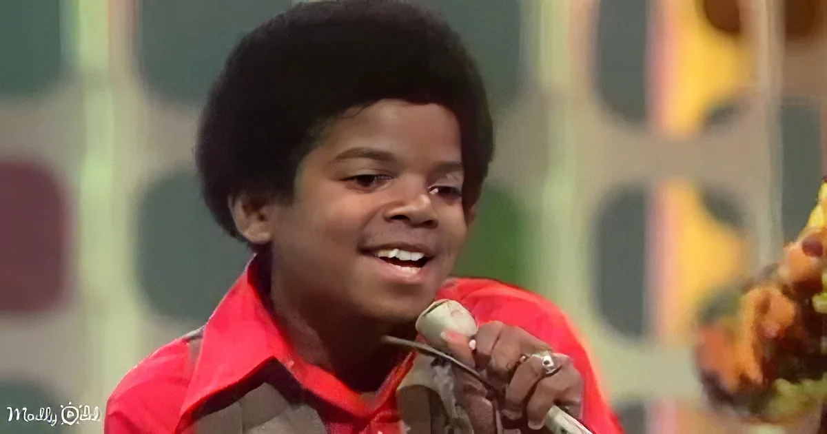 A young boy with a big afro holds a microphone, smiling, wearing a red shirt, against a colorful background.