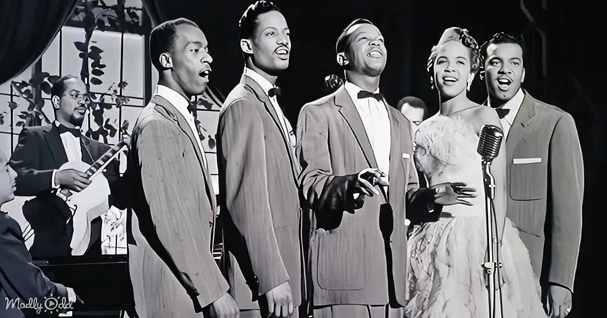 A group of five performers in suits sing on stage, accompanied by a banjo player in the background.