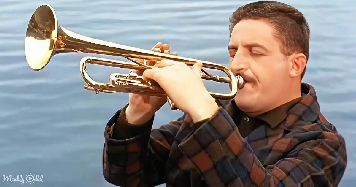 A man in a plaid shirt plays a trumpet with his eyes closed, set against a blue water background.