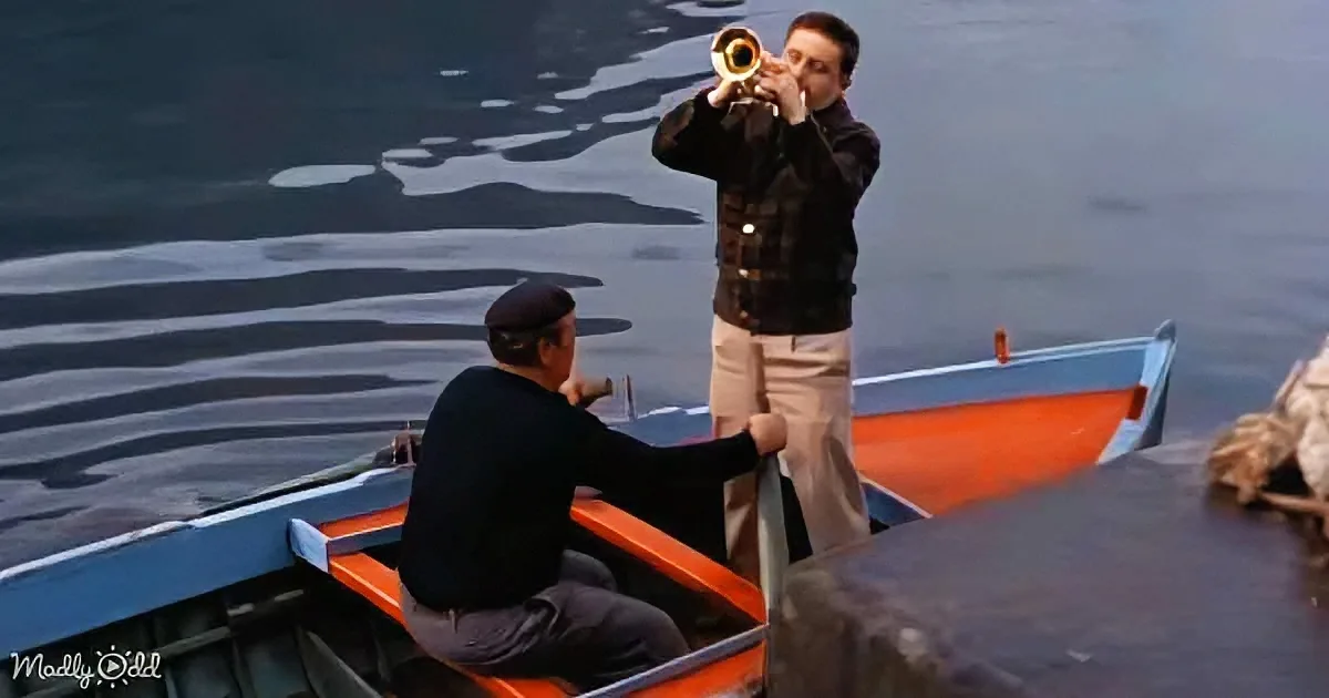 A man plays trumpet on a boat, another man sits nearby, both on a calm body of water.