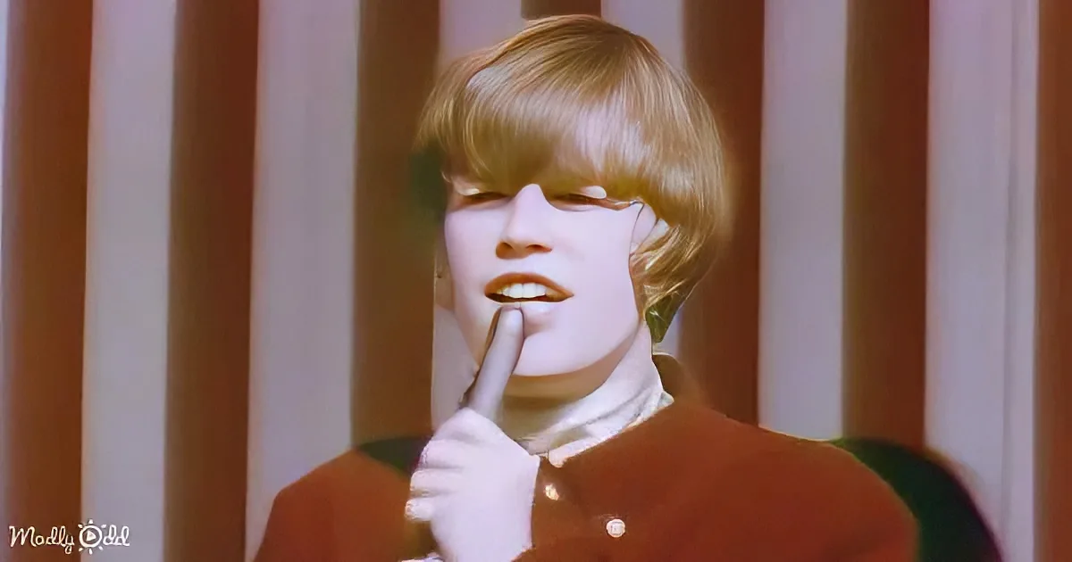A person with blonde hair, wearing a brown jacket and white gloves, holds a finger to their lips.