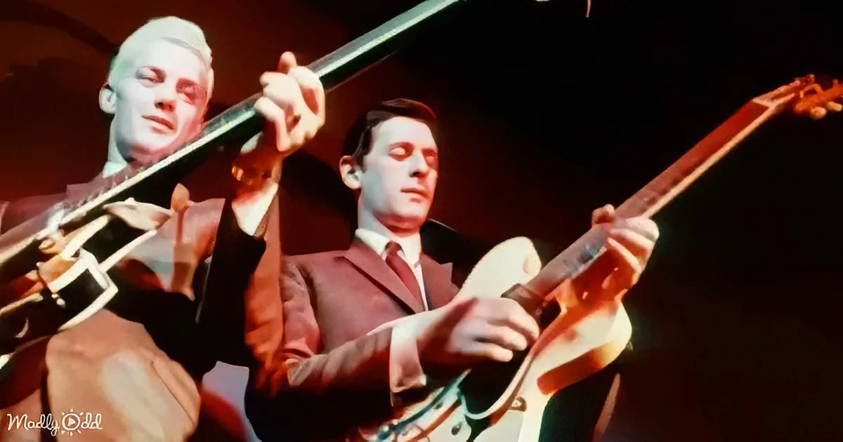 A man in a suit plays a guitar on stage, illuminated by warm lighting.