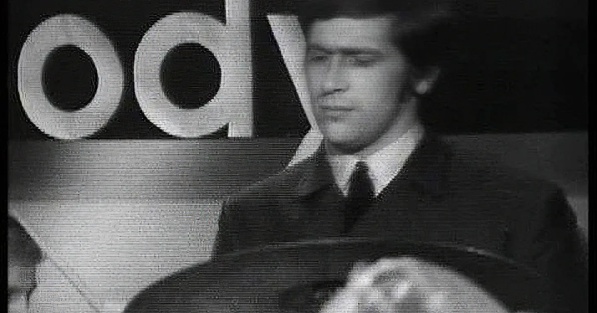 A man with long hair and a suit, captured in a black-and-white close-up, appears to be singing or speaking.