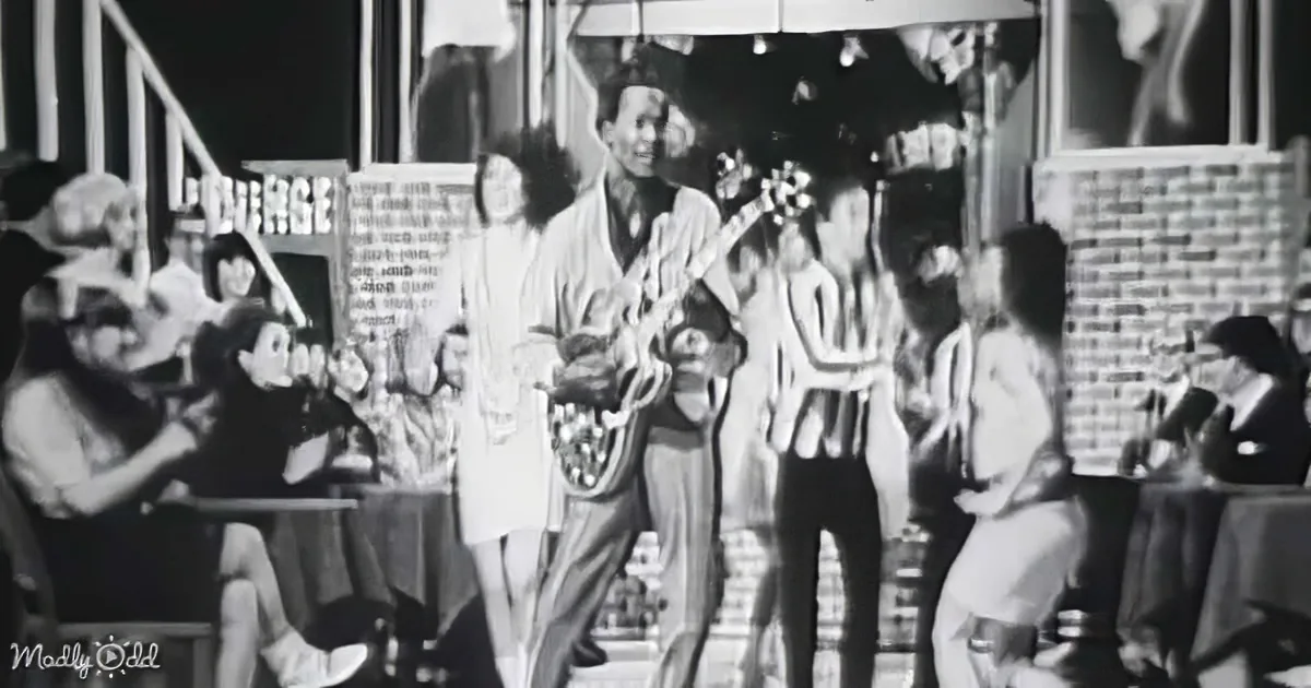 A man plays guitar on stage, surrounded by cheering crowd and brick backdrop.