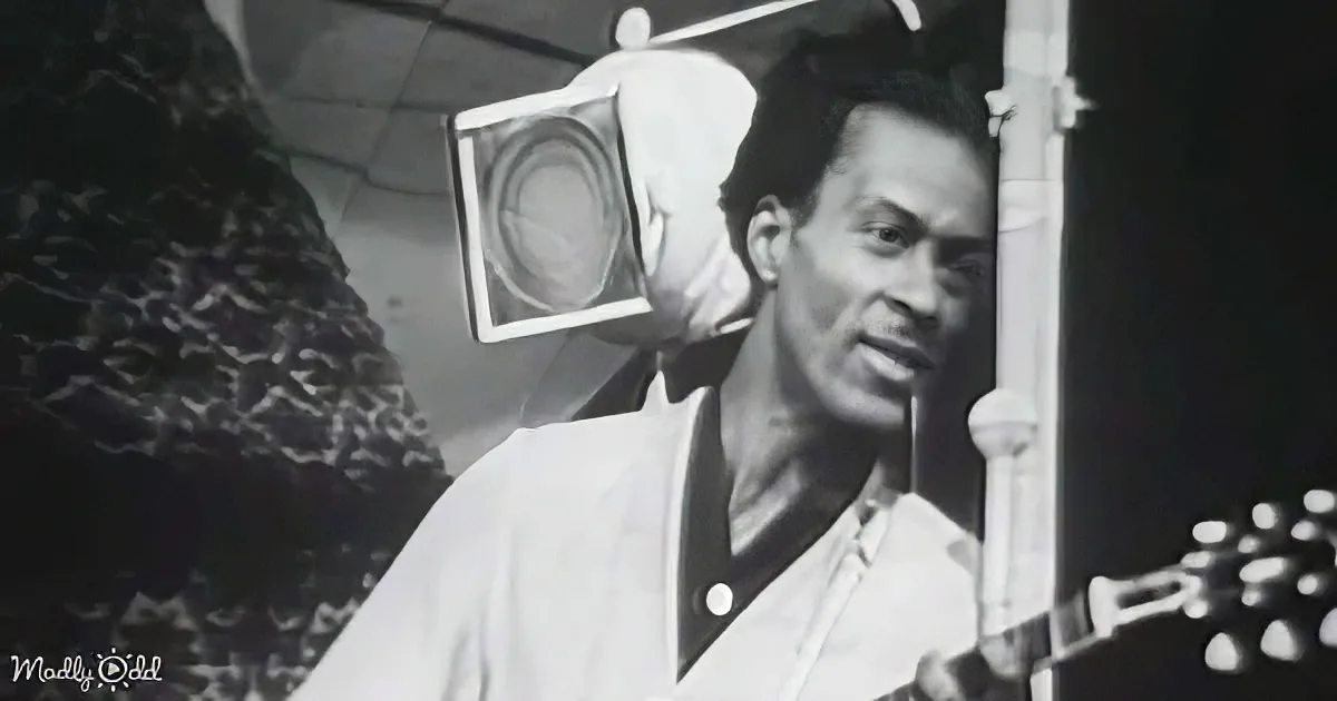 A man in a white shirt is playing a guitar, with a speaker and microphone in the background.