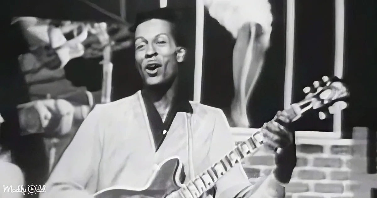 A man in a white shirt plays a guitar while singing, with a brick wall and blurred figures in the background.