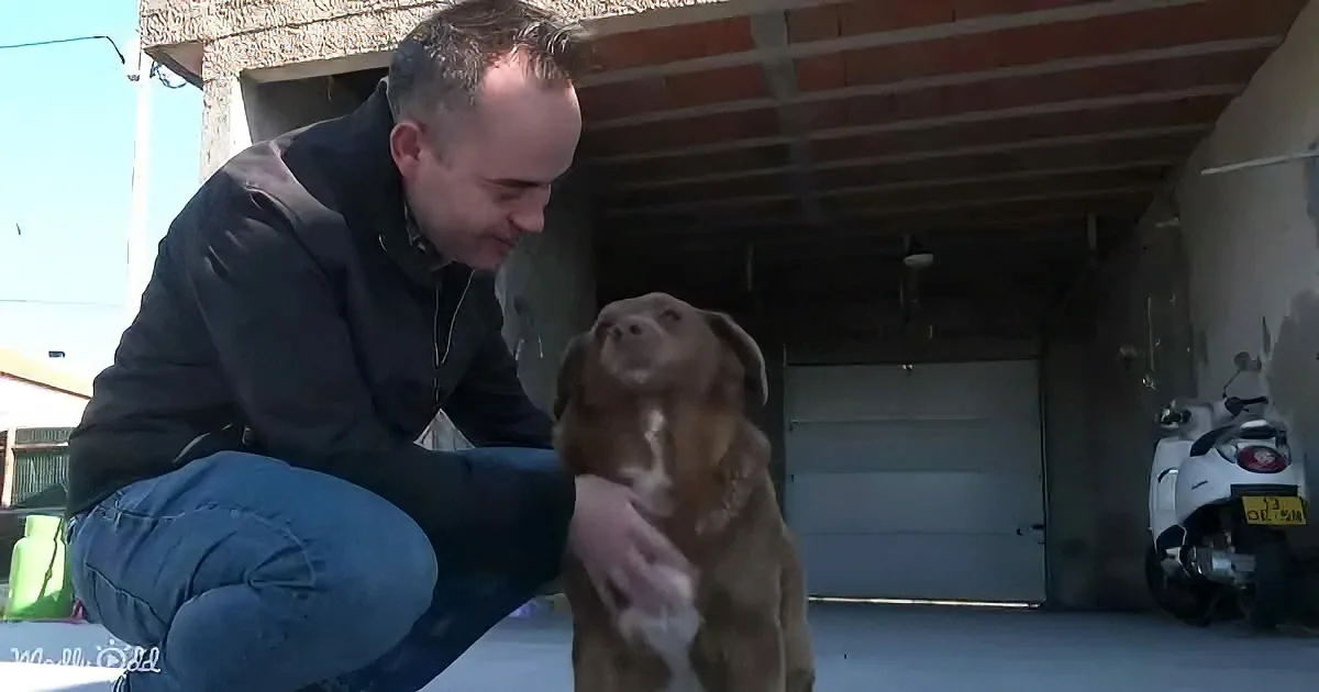 A man kneels to pet a brown dog in front of a garage.