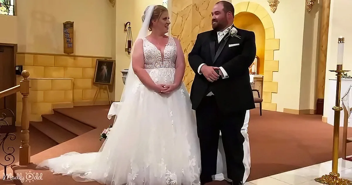 A bride in a white gown and veil stands next to a groom in a black suit, both smiling at each other in a church setting.