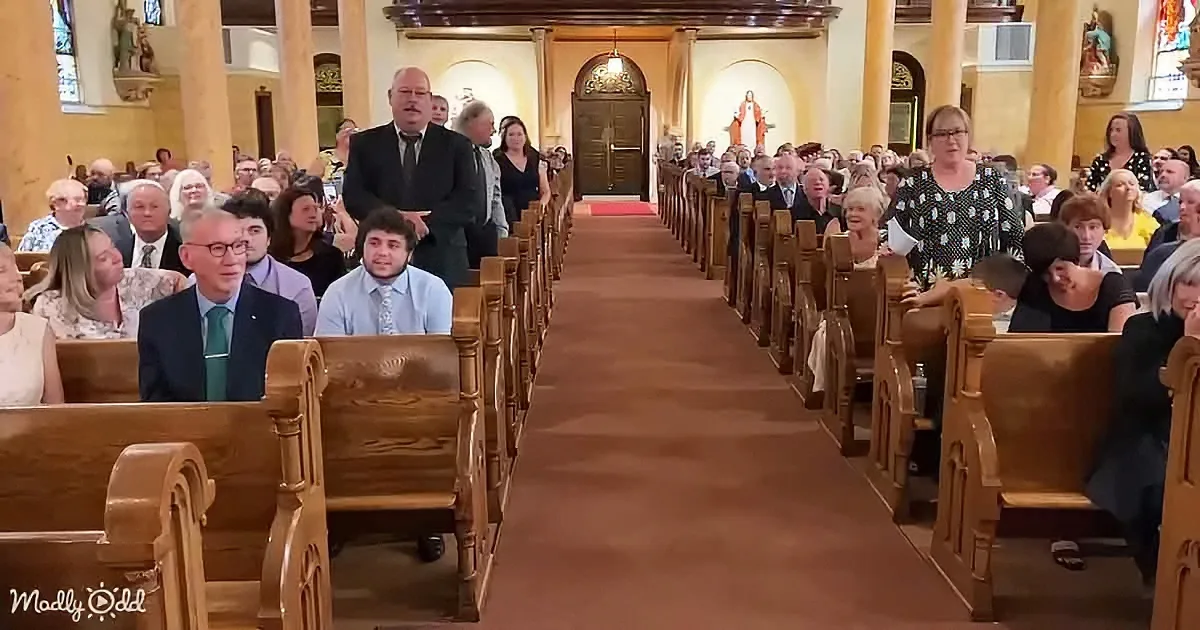 A church interior filled with people seated on wooden pews, facing forward, with stained glass windows and a Jesus statue...