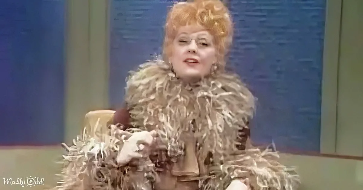 A person in a fluffy, textured outfit sits on a chair, holding a microphone, with a blue background.