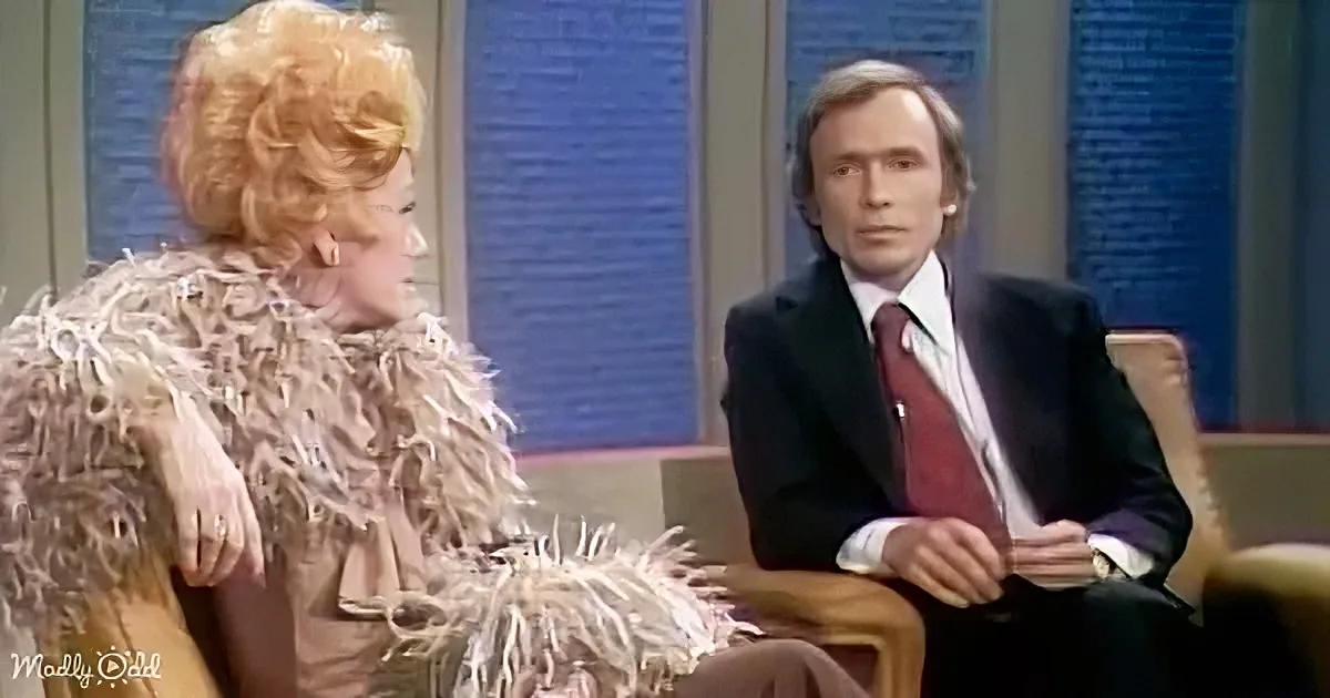 A woman in a feathered dress sits next to a man in a suit on a talk show set.