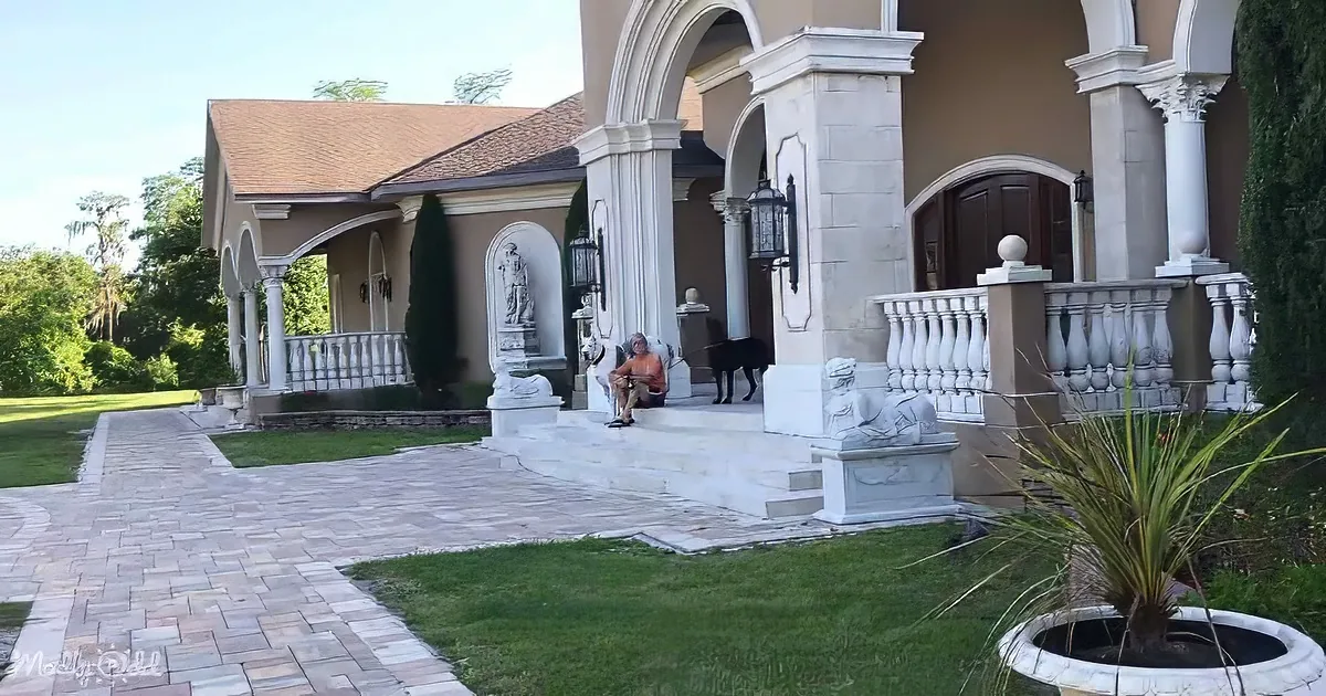 A woman sits on steps near a grand house with statues and a dog, surrounded by greenery and a paved walkway.