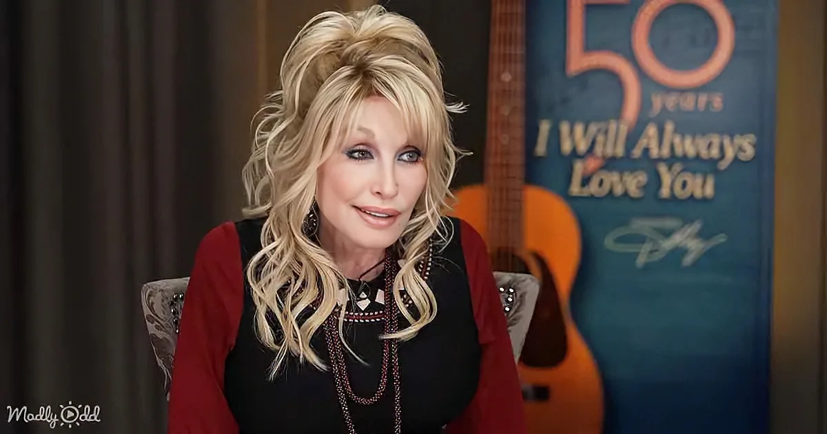 Dolly Parton sits in front of a guitar and a "50 Years I Will Always Love You" sign, wearing a red and black outfit.