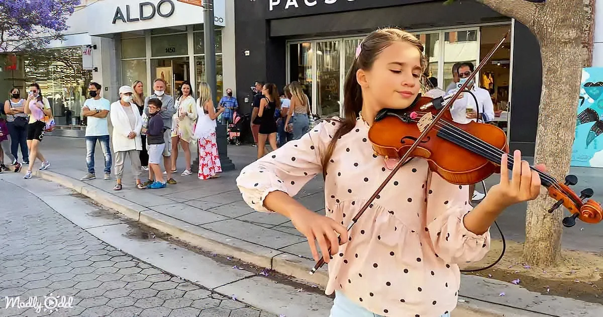 A girl plays violin on sidewalk, people watch behind her, Aldo store in background.