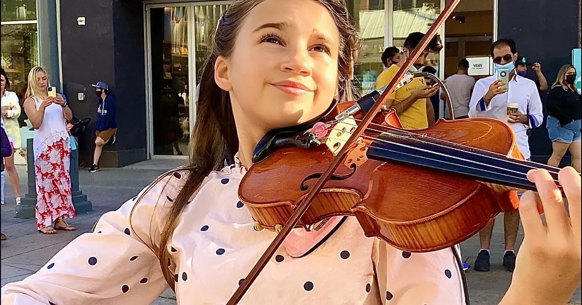 A girl in a polka-dot dress plays the violin on a sidewalk, surrounded by onlookers and a dog.