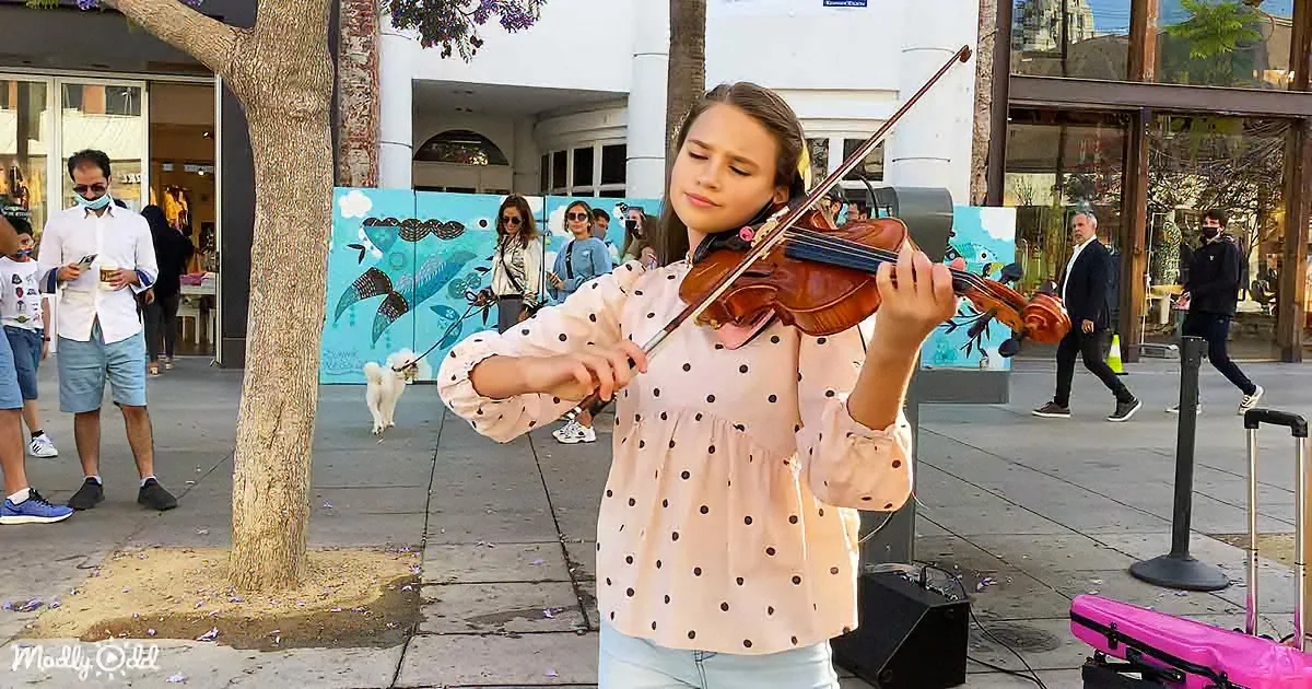 A girl in a polka-dot shirt plays the violin on a city sidewalk, surrounded by pedestrians and colorful murals.