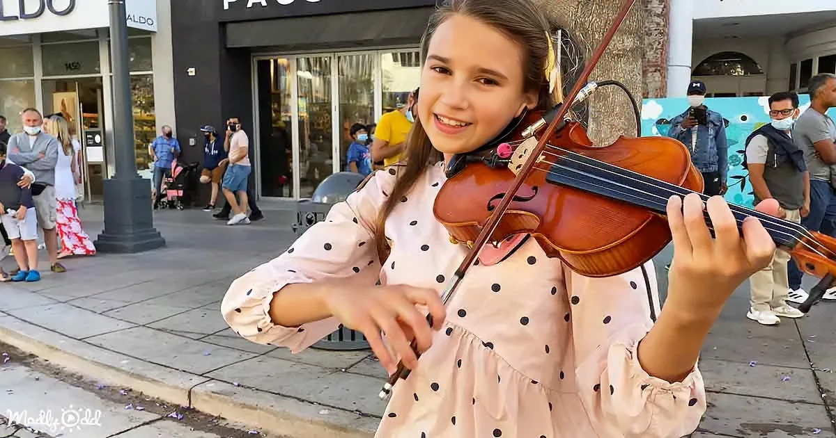 A young girl in a polka-dot dress plays a violin on a city sidewalk, surrounded by onlookers.
