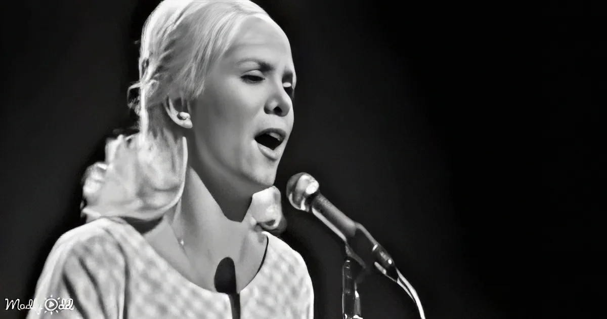 A woman with blonde hair, wearing a patterned top, sings into a microphone against a dark background.