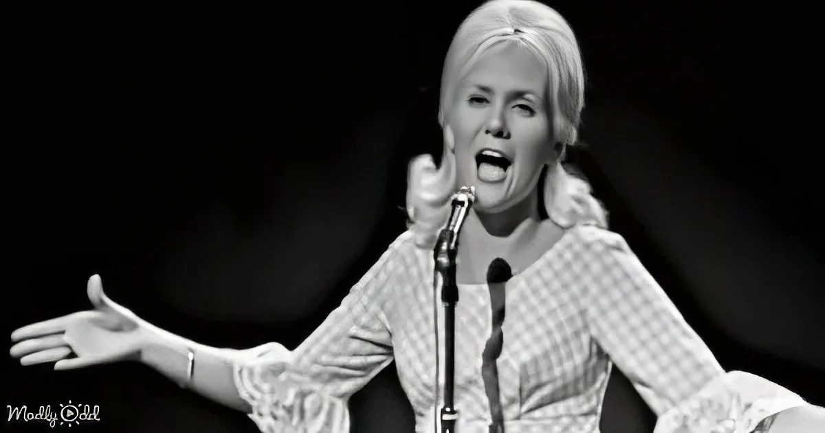 A woman in a checkered shirt sings passionately on stage with her arms outstretched.