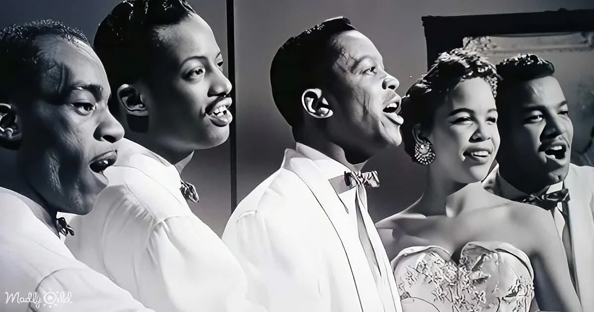 A group of five people in formal attire, singing together, with a woman in a white dress at the front.