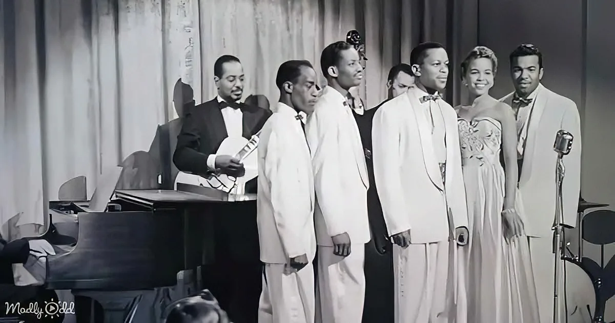 A group of men in white suits and a woman in a dress stand on stage, singing, with a musician playing a stringed...