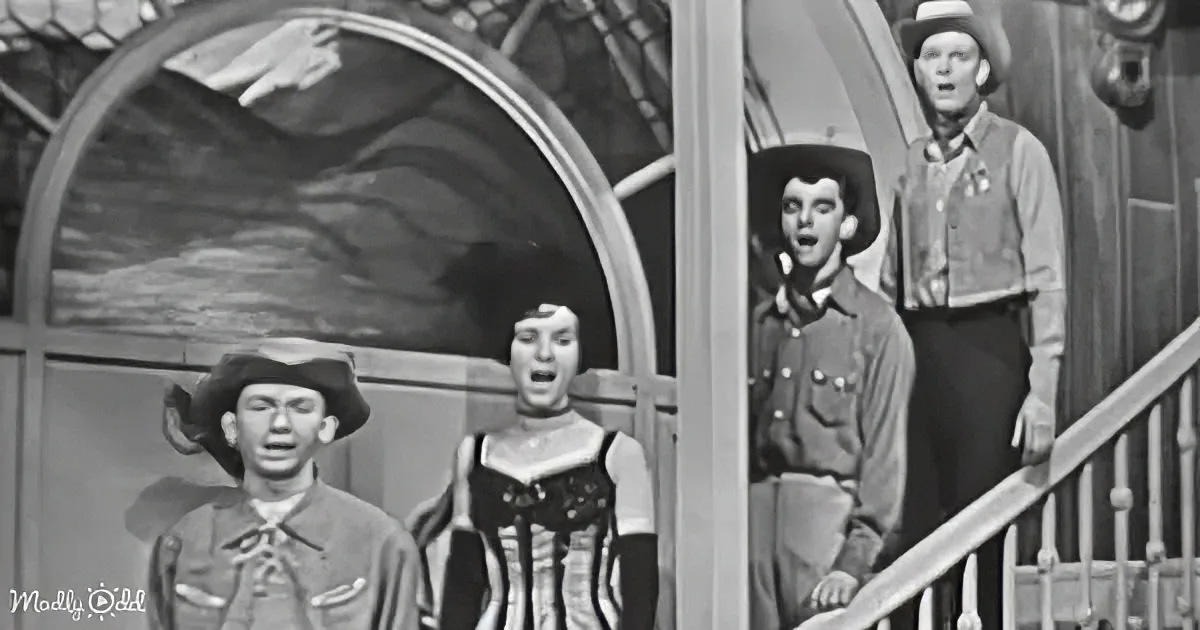 A group of people dressed in Western attire stands in front of a rustic setting, seemingly singing or performing.