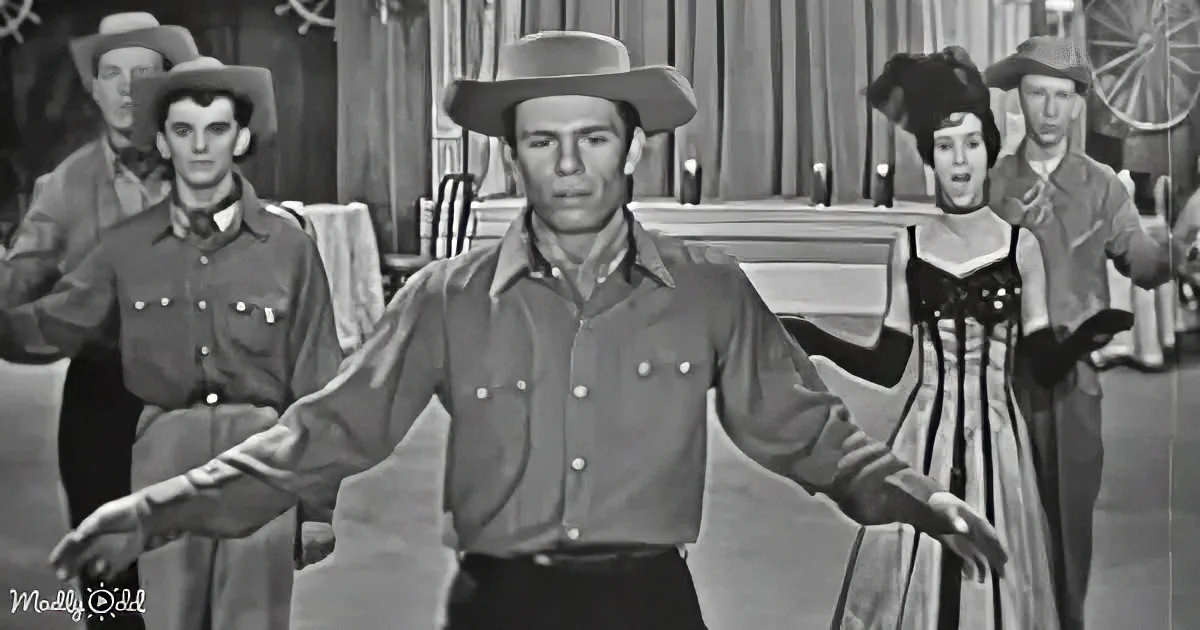 A man in a cowboy hat and shirt dances in front of a group in a Western-style setting.