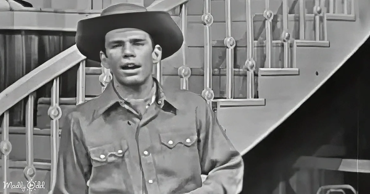 A man in a cowboy hat and shirt stands in front of a staircase, looking to the side.