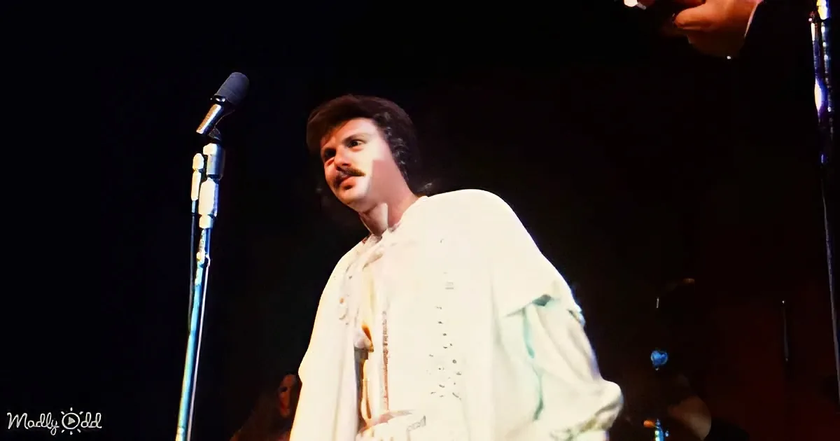 A man in a white outfit stands on stage with a microphone, looking to the side.