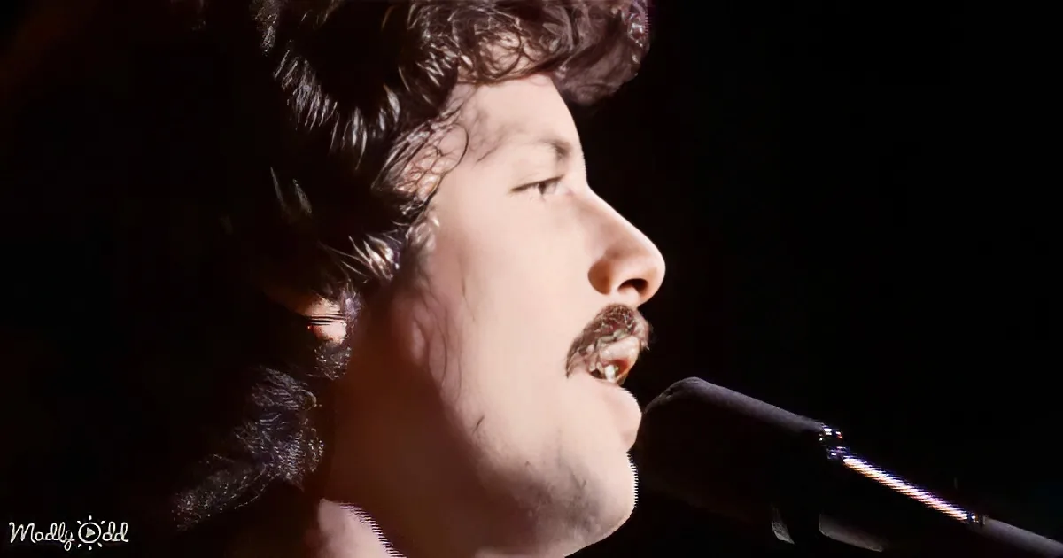 A person with curly hair and a mustache sings into a microphone, eyes closed, in a dimly lit setting.