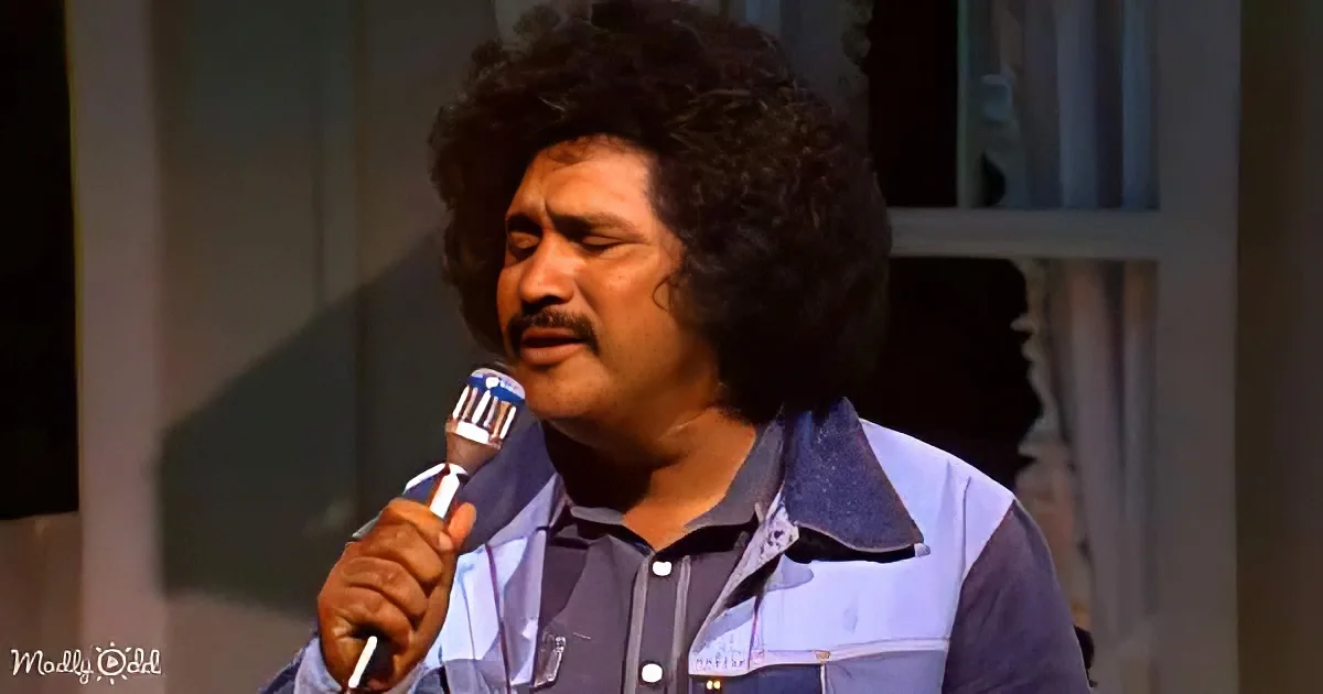 A man with curly hair and a mustache holds a microphone, wearing a blue shirt, in a dimly lit setting.