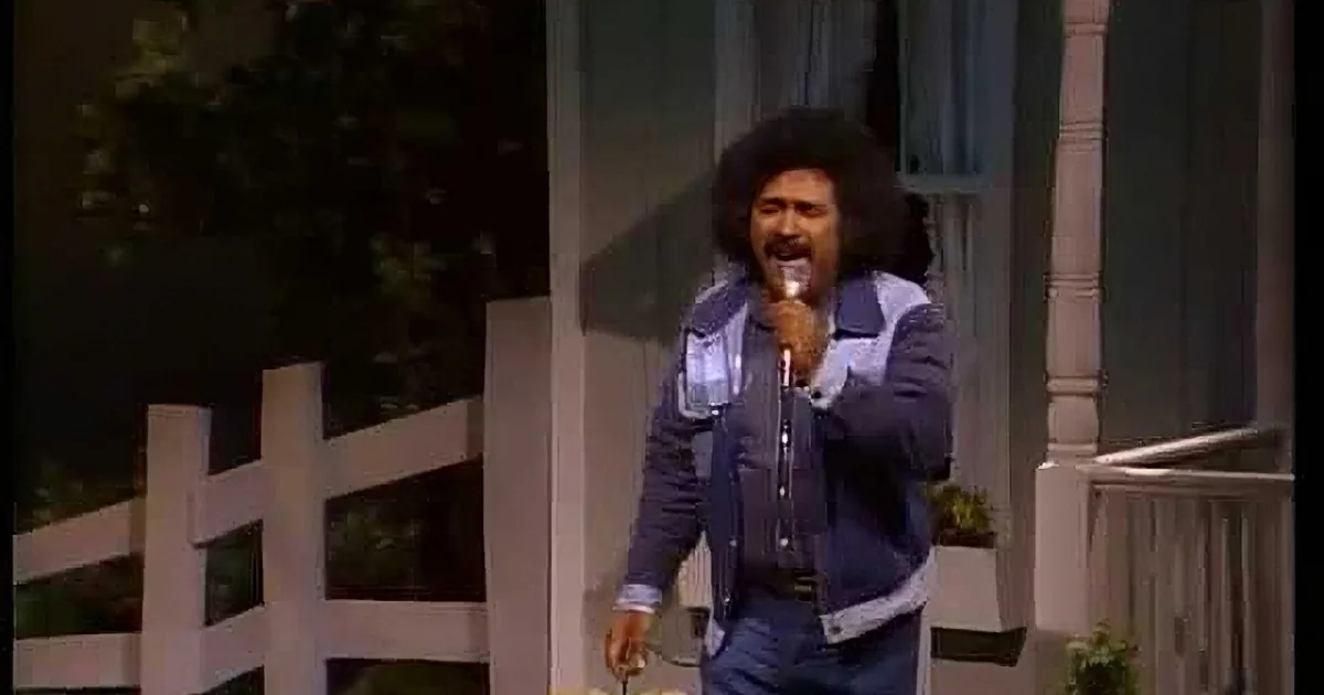 A man with curly hair and a mustache sings into a microphone, wearing a denim vest over a shirt.