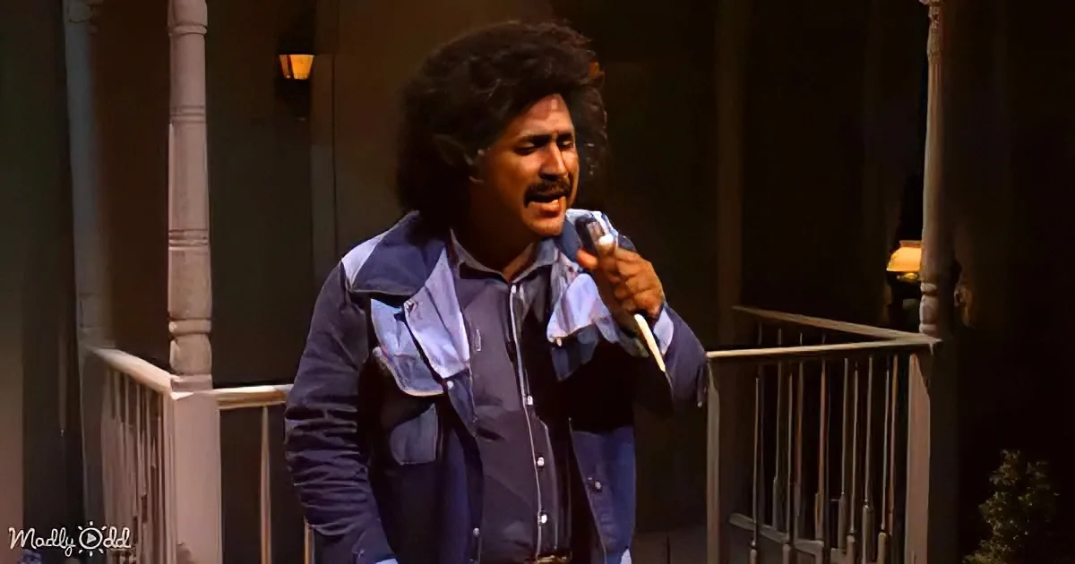 A man with curly hair and a mustache sings into a microphone on a porch.