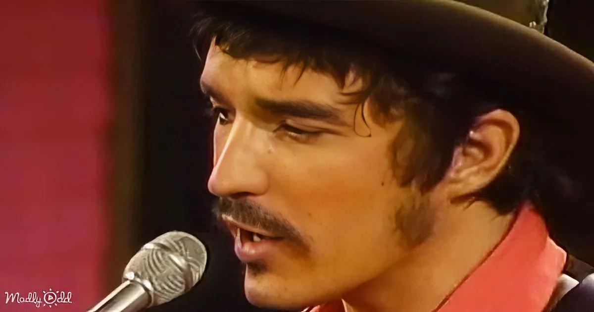A man in a cowboy hat sings into a microphone, wearing a red shirt.