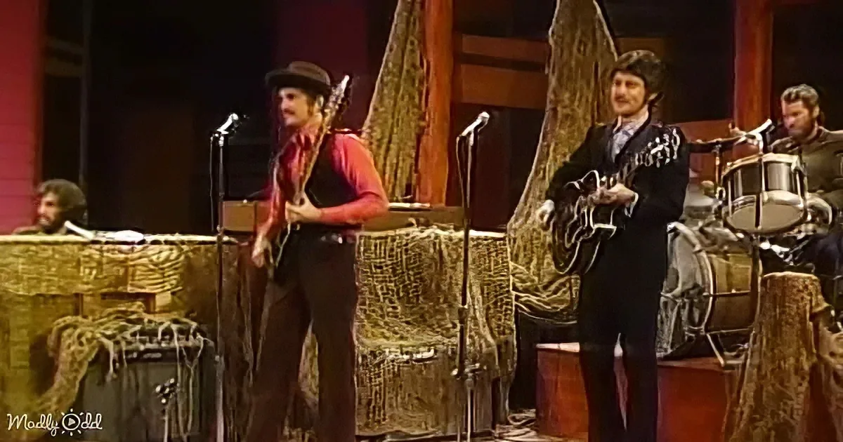 A man in a red shirt and hat plays guitar on stage with a band.