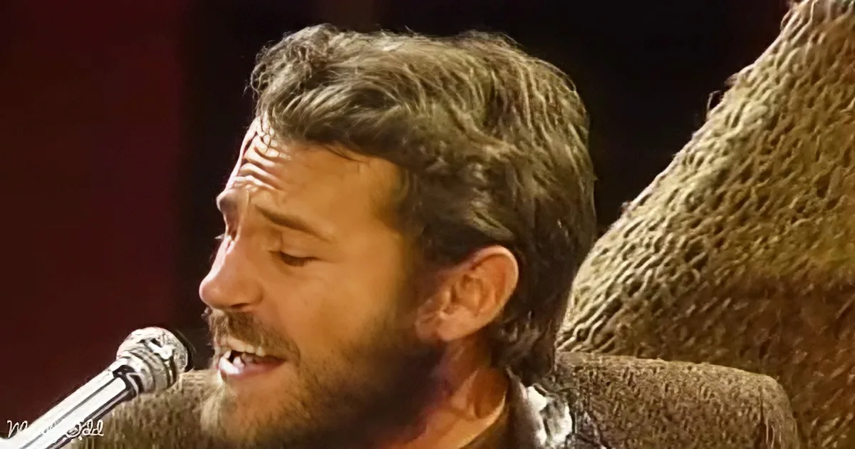A man with short, light brown hair and a beard sings into a microphone, wearing a textured brown jacket.