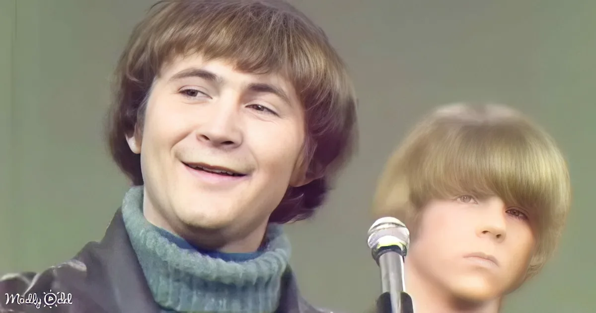 A smiling person with shoulder-length hair, wearing a turtleneck, stands in front of a microphone, with another person in...