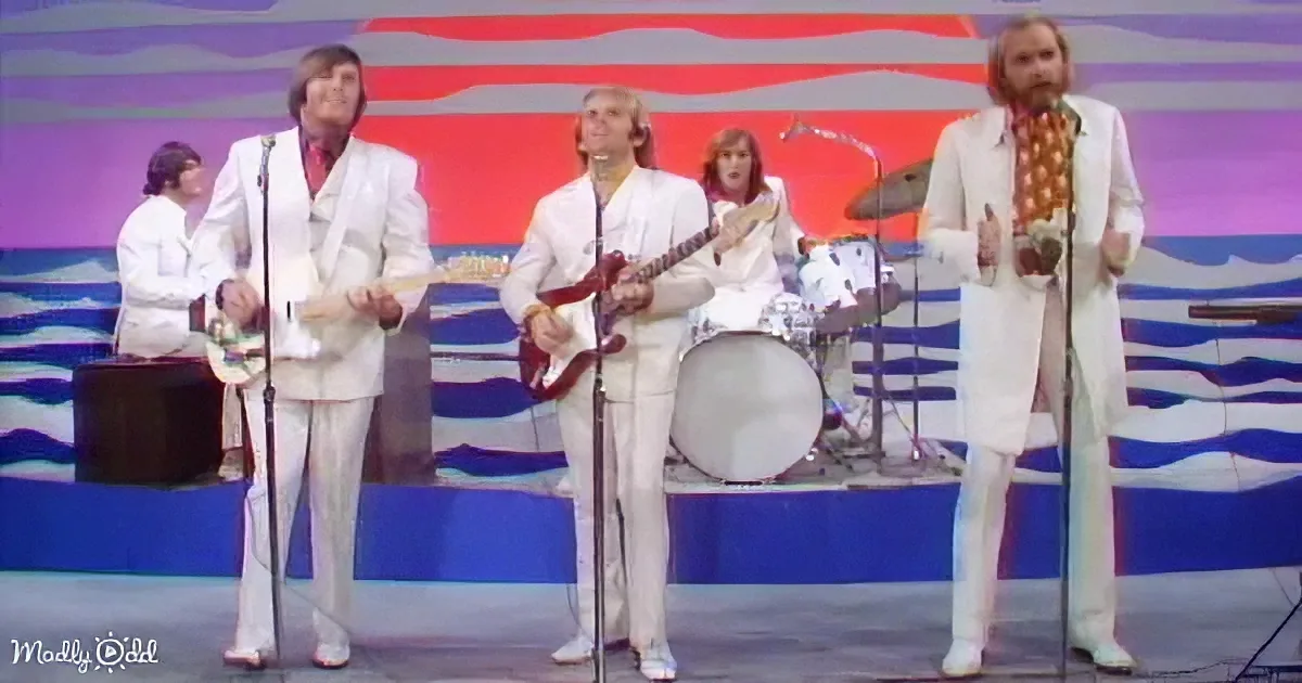 A band in white suits performs on stage with a colorful backdrop, holding microphones and instruments.