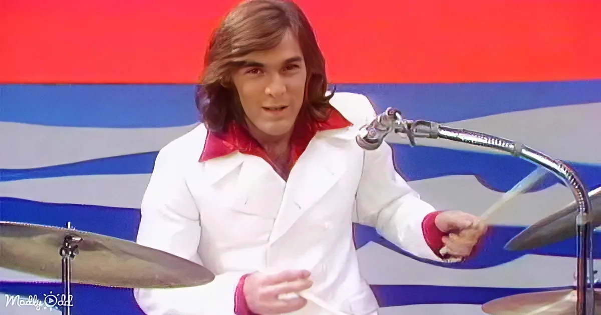A person in a white shirt with red accents plays drums in front of a red and blue striped background.